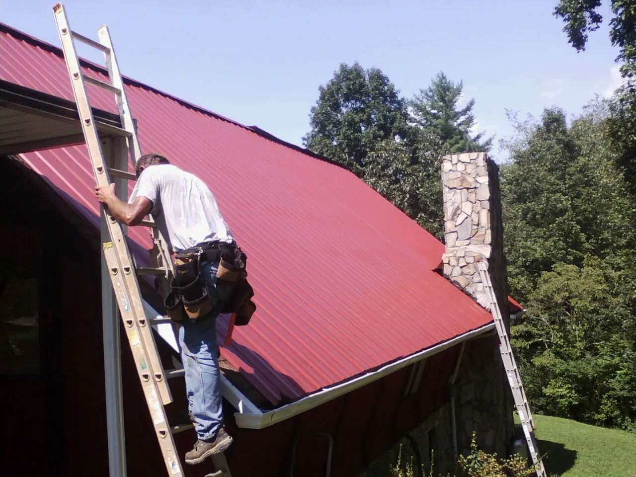 Roofer on a ladder beside a red metal roof and stone chimney — Soffit & Fascia Repair by Roofing Brothers in Lenoir City